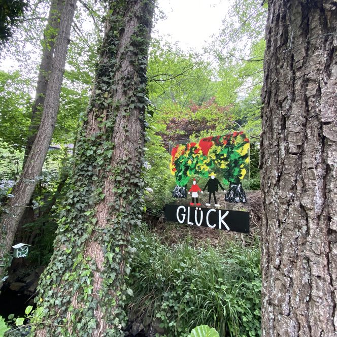 <p>Waldlandschaft mit buntem Kunstwerk und Wort "GLÜCK", umgeben von üppigem Grün und Bäumen.</p>