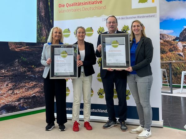 Drei Frauen und ein Mann stehen vor einer Leinwand und halten zwei Zertifikate in den Händen.