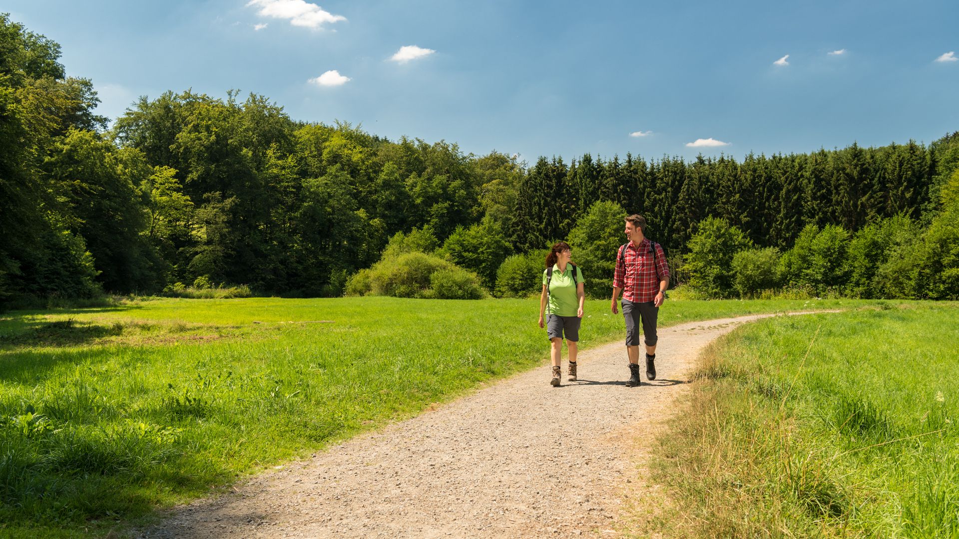 Ein Paar wandert über den Eifgenbachweg, links und rechts vom Weg saftig grünes Gras, im Hintergrund ein Wald und blauer Himmel.
