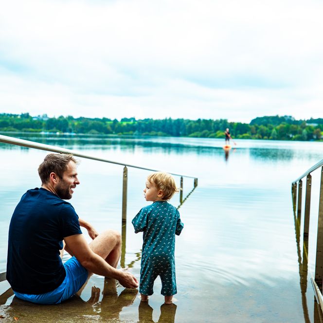 Ein Vater und ein Kind genießen gemeinsam Zeit am Rand eines ruhigen Sees mit bewaldetem Ufer.
