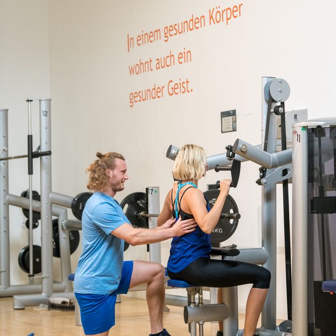 Ein Trainer unterstützt eine Frau beim Training an einem Fitnessgerät in einem hellen Studio.