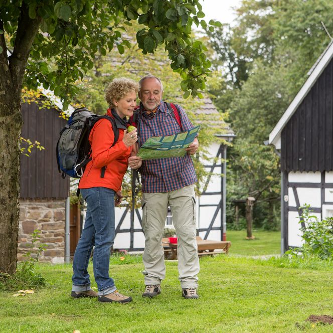 Zwei Senioren mit Rucksack lesen eine Karte vor traditionellen Fachwerkhäusern im Grünen.