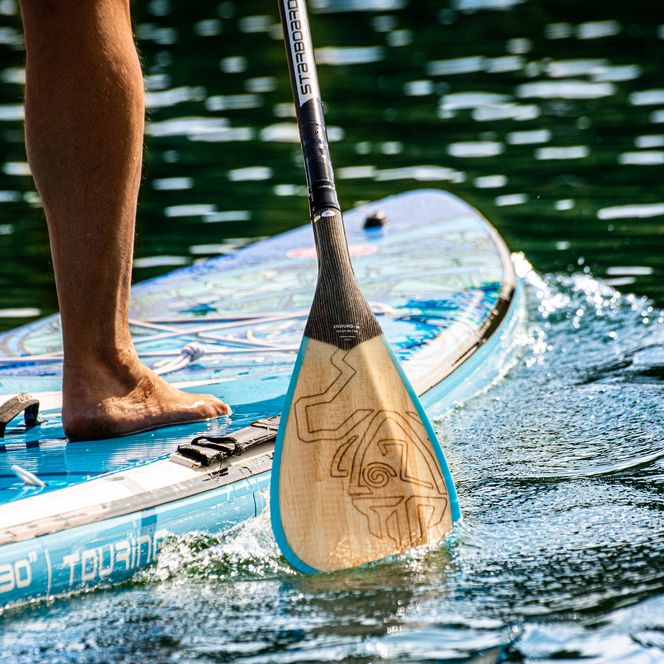 Nahaufnahme eines Paddels und Beins auf einem SUP-Board auf glitzerndem Wasser.