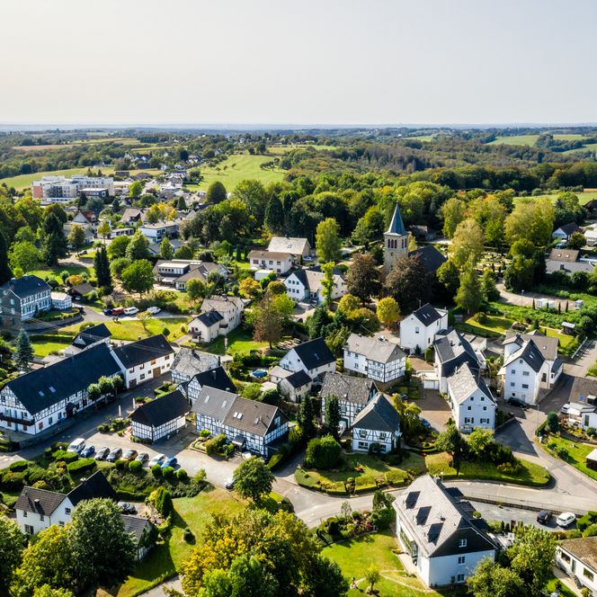 Luftaufnahme eines idyllischen Dorfes mit Kirche, umgeben von bewaldeten Hügeln und grünen Landschaften.