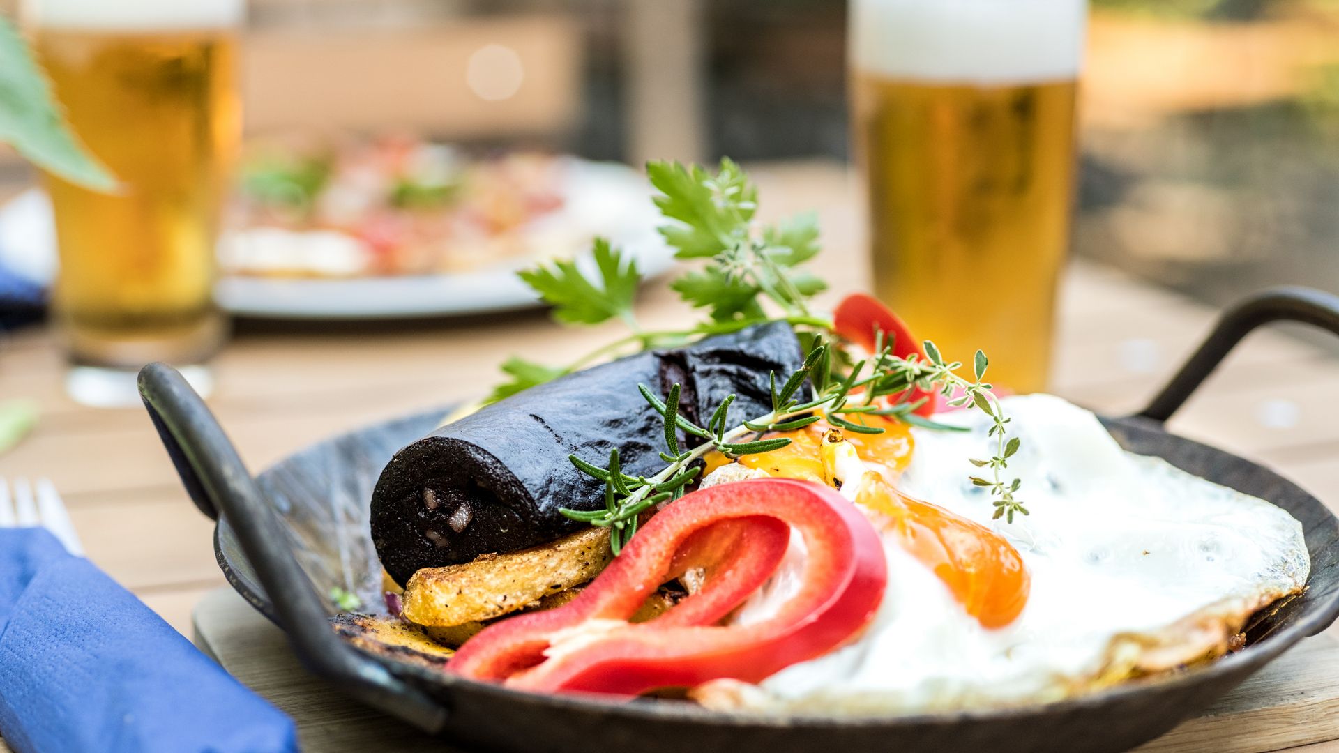 Im Vordergrund ist ein Pfännchen mit Bratkartoffeln, Blutwurst und einem Spiegelei, dekoriert mit Paprika auf einem Holztisch. Daneben steht ein Glas Bier. Im Hintergruns ist unscharf ein weiteres Gericht und ein weiteres Glas Bier sichtbar. 