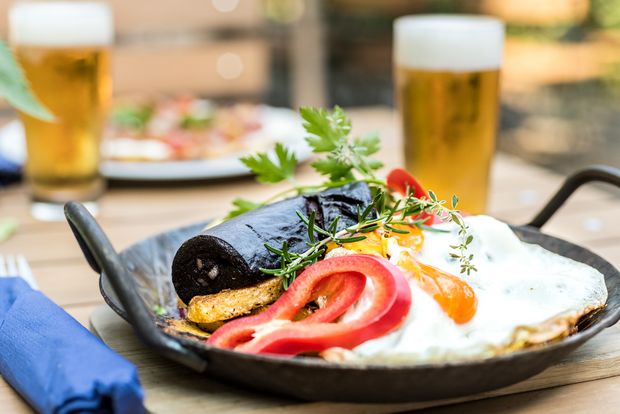 Im Vordergrund ist ein Pfännchen mit Bratkartoffeln, Blutwurst und einem Spiegelei, dekoriert mit Paprika auf einem Holztisch. Daneben steht ein Glas Bier. Im Hintergruns ist unscharf ein weiteres Gericht und ein weiteres Glas Bier sichtbar. 