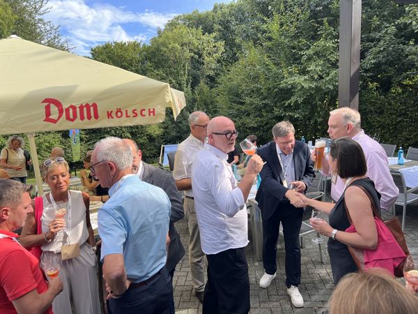 Teilnehmer einer Veranstaltung stehen auf einer Terrasse unter einem Sonnenschirm und unterhalten sich.