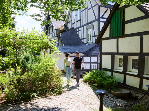 Ein Pärchen spaziert vor mehreren Fachwerkhäusern bei sonnigem Wetter. Links ist ein grüner Garten zu sehen.