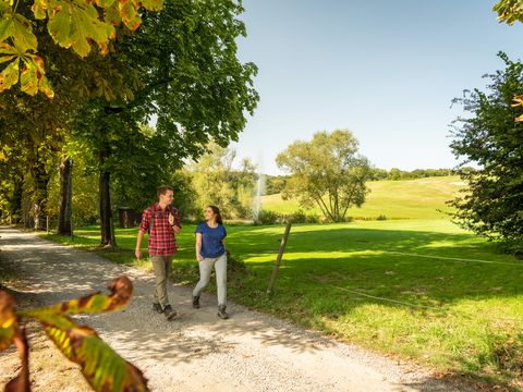 Ein junges Pärchen wandert auf einem flachen Wanderweg. Sie schauen sich an. Im Hintergrund ist ein Golfplatz und ein Springbrunnen zu sehen.