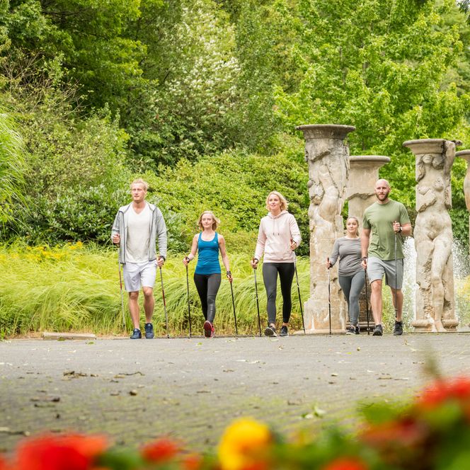 <p>Fünf Personen wandern in sommerlicher Kleidung durch einen grünen Park mit antiken Säulen.</p>