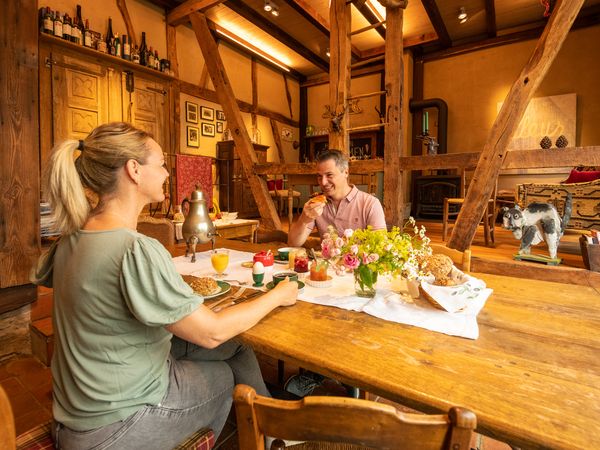 Ein Pärchen sitzt sich an einem gedeckten Frühstückstisch gegenüber in einem gemütlichen Holzhaus. 