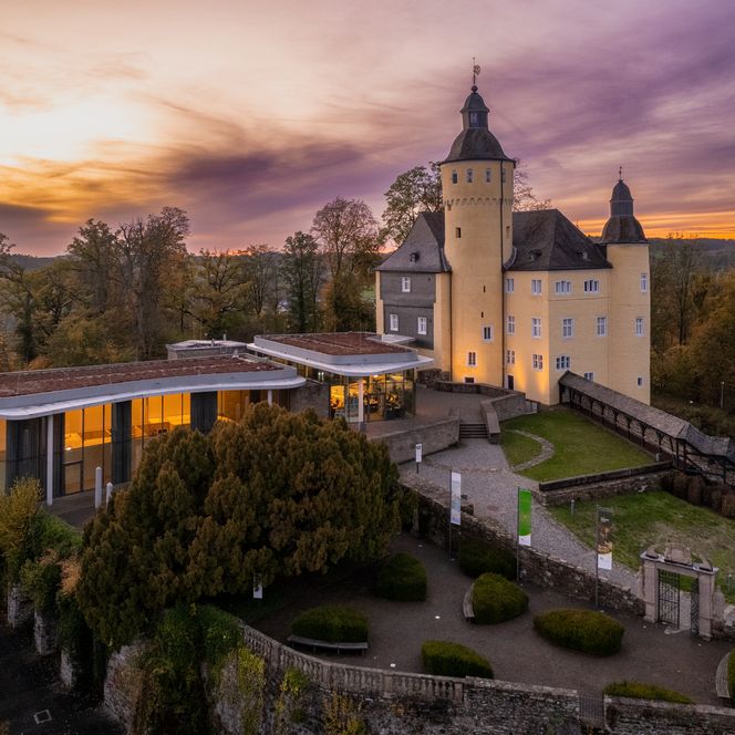 <p>Schloss Homburg bei Sonnenuntergang, umgeben von Bäumen und einem modernen Anbau, im Herbst.</p>