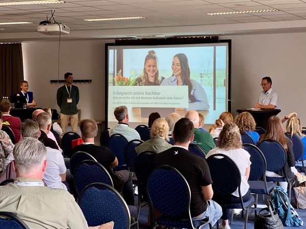 Teilnehmer einer Veranstaltung sitzen in einem Tagungsraum vor einer Leinwand. Sie sind von hinten zu sehen.
