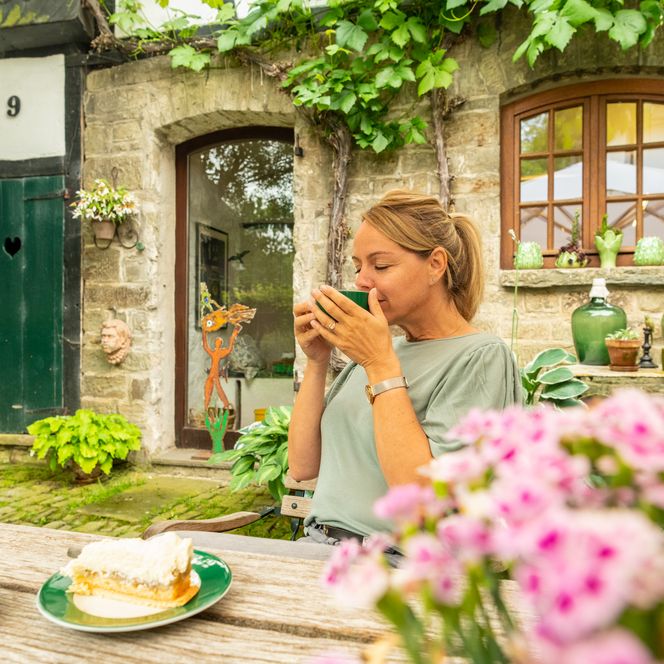Eine Frau genießt Kaffee in einem idyllischen Garten mit Blumen und einem Stück Kuchen.