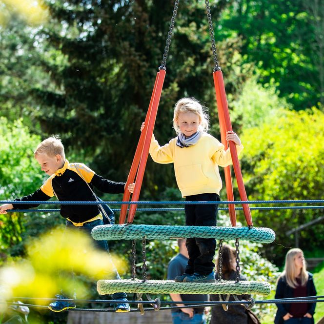 <p>Zwei Kinder spielen fröhlich auf einem Seilspielgerät in einem sonnigen Park.</p>