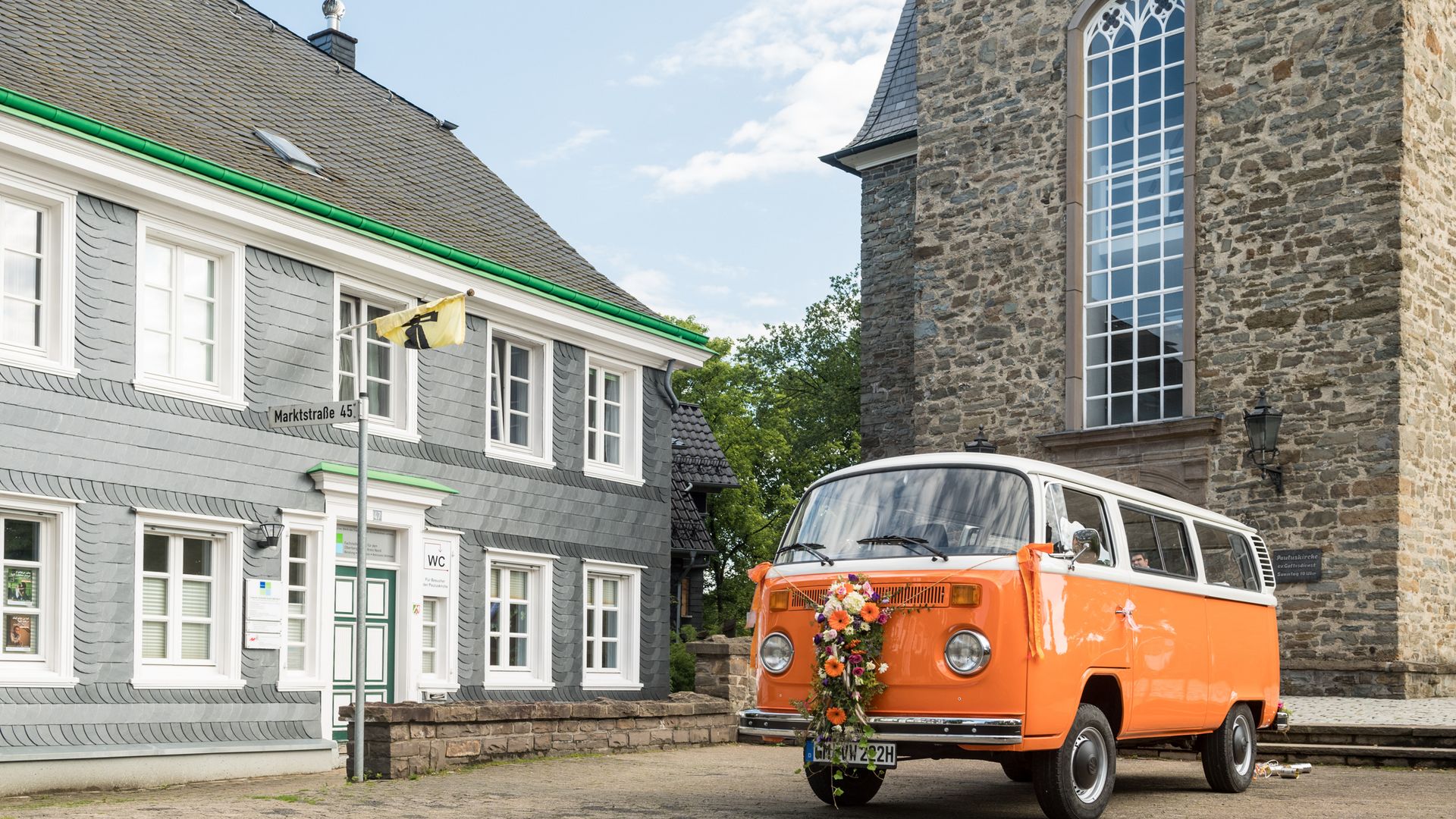 ein orangefarbener Oldtimer steht vor einer gemauerten Wand und einem Schieferhaus.