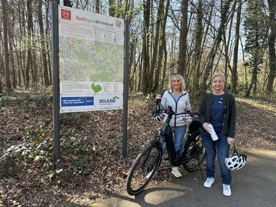 Zwei Radfahrerinnen stehen vor einer Infotafel im Wald. Eine hält ein Fahrrad, die anderen einen Helm und eine Trinkflasche.