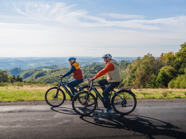 Ein Mann in eine Frau fahren auf Fahrrädern. Im Hintergrund ist bergische Landschaft zu sehen. 