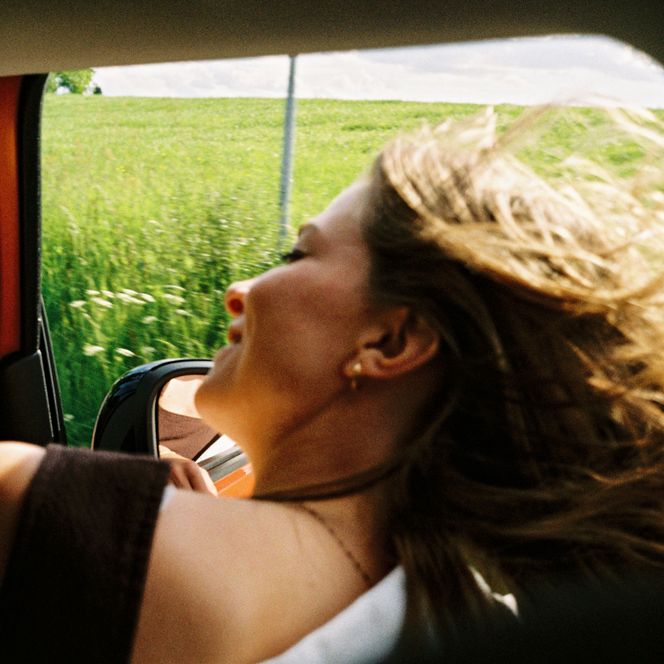 Eine Frau mit blonden Haaren hält ihren Kopf aus dem Fenster eines fahrenden Vans in den Wind.