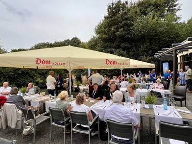 Teilnehmer einer Veranstaltung sitzen auf einer Terrasse unter einem großen gelben Sonnenschirm. 
