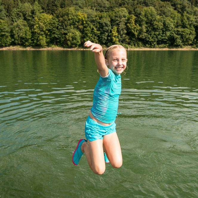 <p>Ein fröhliches Kind springt in einem blauen Badeanzug in einen See, umgeben von Wald.</p>