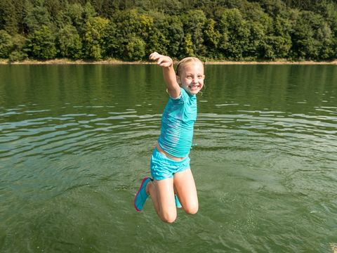 Mädchen springt mit hochgezogenen Beinen und ausgestreckten Armen in den Badesee