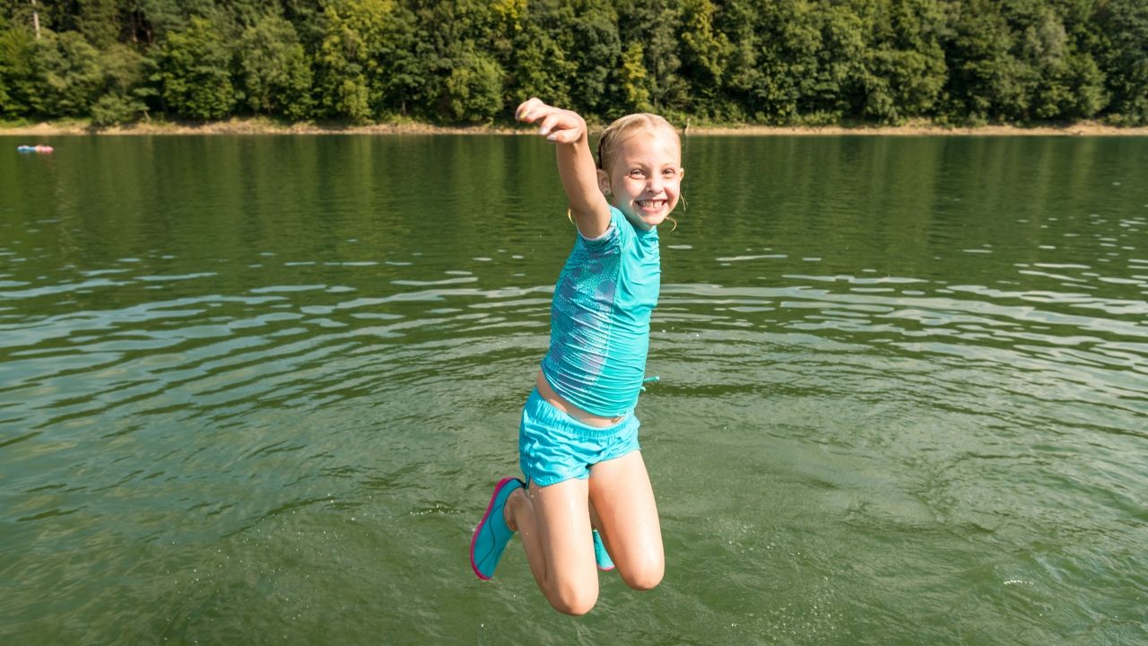 Mädchen springt mit hochgezogenen Beinen und ausgestreckten Armen in den Badesee