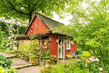Kleines, rotes Gartenhaus umgeben von üppigem Grün und bunten Blumen im Dorf Much.