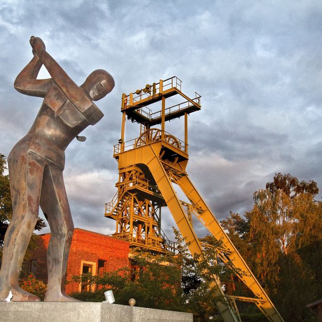 Industriedenkmal mit gelbem Förderturm, flankiert von einer Bergarbeiterstatue und umgeben von Bäumen.
