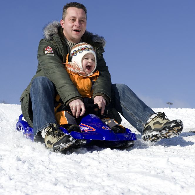 Mann und Kind rodeln fröhlich im Schnee auf einem blauen Schlitten unter klarem, blauem Himmel.