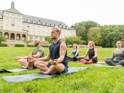 Eine Gruppe mit 3 Frauen und 2 Männern sitzt auf Yogamatten auf einer Wiese vor einem Hotelgebäude und meditiert. 