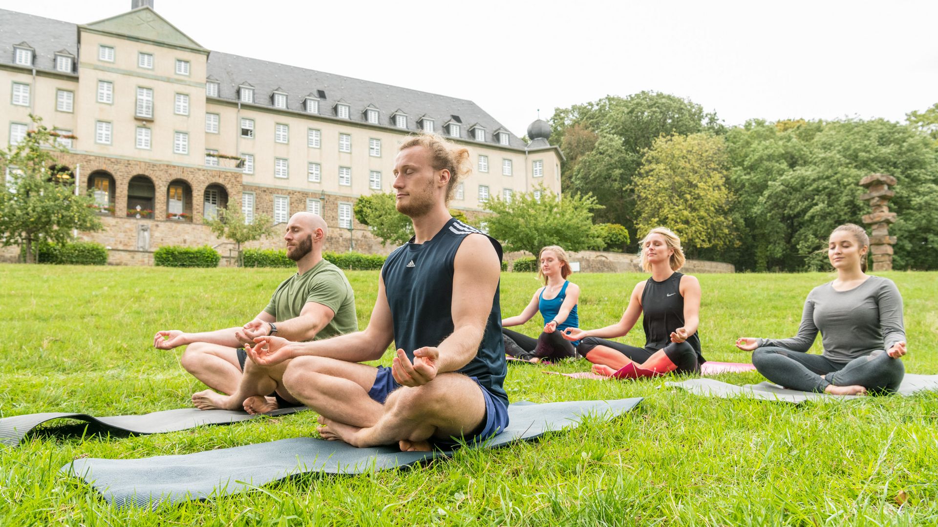 Yoga-Veranstaltung mit drei Frauen und zwei Männern im Schneidersitz auf Yoga-Matten auf einer Wiese, im Hintergrund das Kardinal Schulte Haus