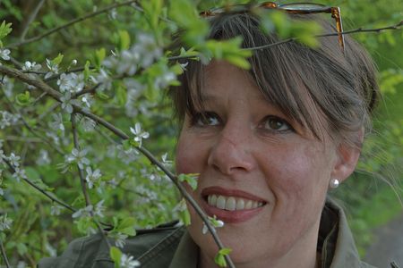 Frau mit braunem Haar lächelt durch einen blühenden Strauch. Im Hintergrund grünes Laub.