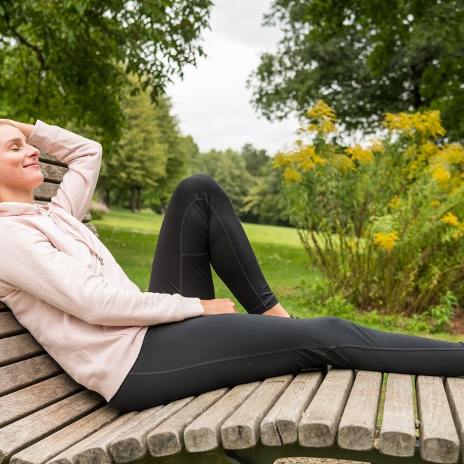 <p>Frau entspannt auf einer geschwungenen Holzbank im Park, umgeben von grünen Bäumen und Blumen.</p>