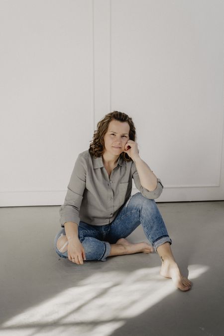 Eine Frau in Jeans und grauer Bluse sitzt barfuß entspannt auf dem Boden in hellem Raum.