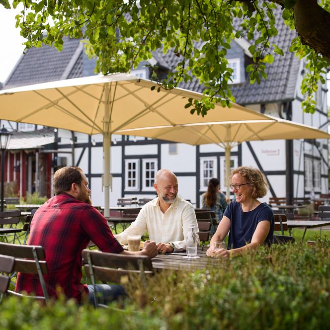 Gemütlicher Biergarten mit Menschen an Holztischen, umgeben von grünen Bäumen und Fachwerkhaus im Hintergrund.