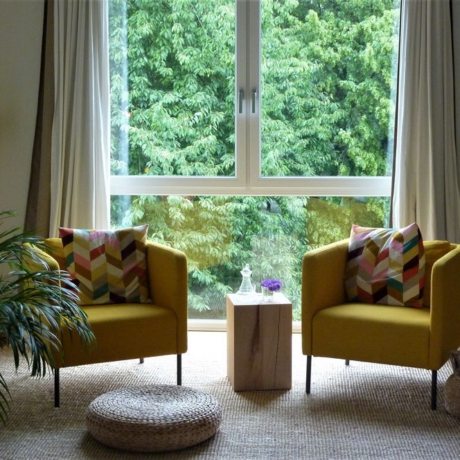 Zwei gelbe Sessel mit bunten Kissen stehen vor einem großen Fenster mit Blick auf grüne Bäume.