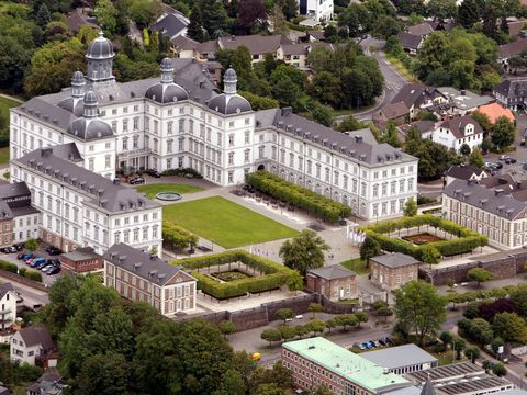 Luftaufnahme auf ein weißes U-förmig angelegtes Schloss mit Rasen im Innenhof in städtischer Bebauung. 