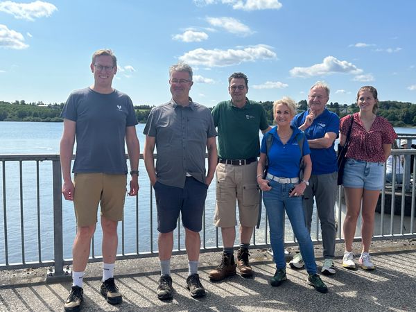 Sechs Menschen stehen vor einem Geländer am Ufer einer Talsperre.