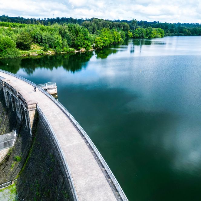 <p>Große Staumauer aus Stein mit Gehweg, umgeben von Wald und Wasseroberfläche in Marienheide.</p>
