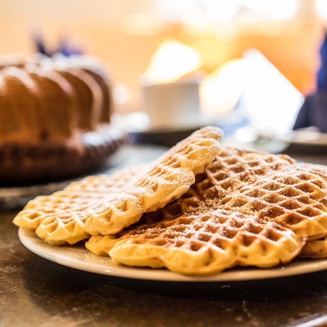 Waffeln mit Puderzucker auf einem Teller; im Hintergrund Kuchen und Kaffeetassen.