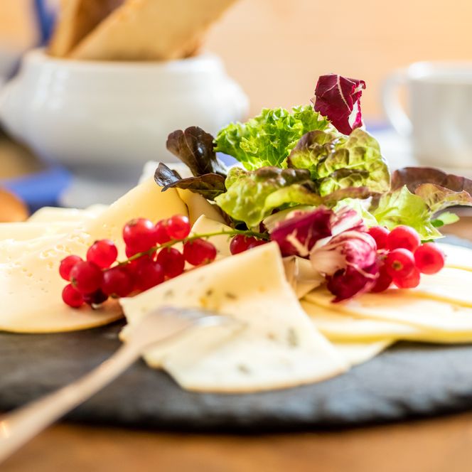 Käsescheiben mit Johannisbeeren und Salat auf Schieferplatte, Besteck und Tasse im Hintergrund.