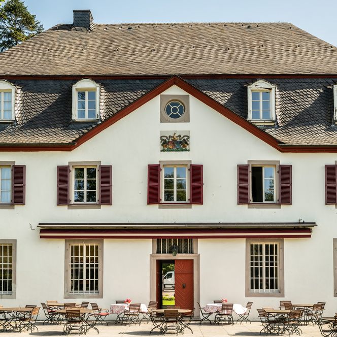 <p>Historisches Gebäude mit Caféterrasse und roten Fensterläden, umgeben von grünen Bäumen.</p>