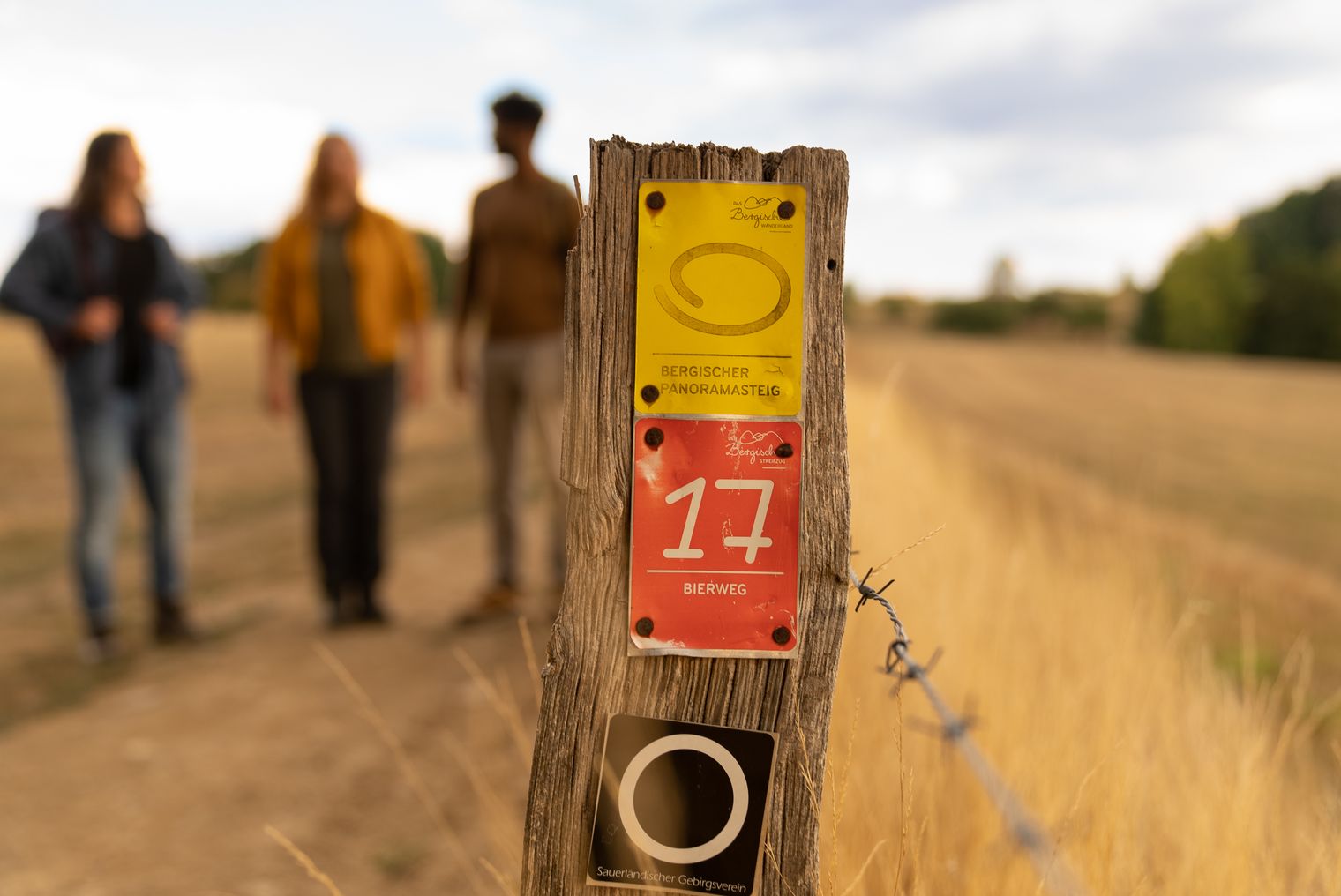 Nahaufnahme eines Holzzauns mit Wanderwegschildern; unscharfe Wanderer im Hintergrund auf einem Pfad.