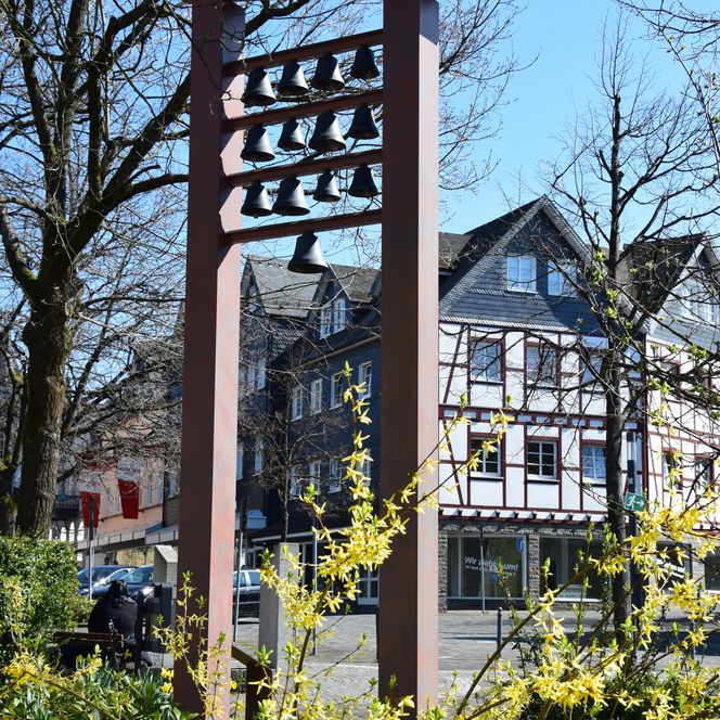 <p>Blumen und Glockenspiel im Vordergrund, Fachwerkhäuser im Hintergrund bei sonnigem Wetter.</p>