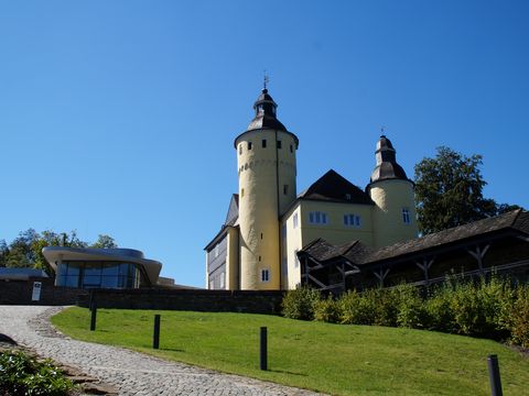 Ein gelbes Schloss steht auf einem Hügel. Im Vordergrund sind eine Wiese und ein Teil einer Straße zu sehen.