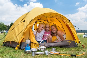 <p>Drei fröhliche Kinder in einem gelben Zelt auf einer Wiese, umgeben von Campingausrüstung.</p>