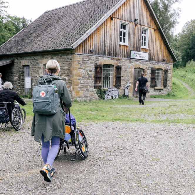 <p>Menschen in Rollstühlen besuchen ein historisches Gebäude auf einem klaren Weg in ländlicher Umgebung.</p>