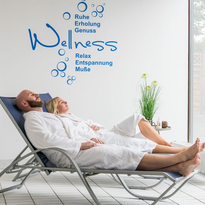 Entspannendes Paar im Wellnessbereich auf Liegestühlen, eingehüllt in Bademäntel, vor weißer Wand.