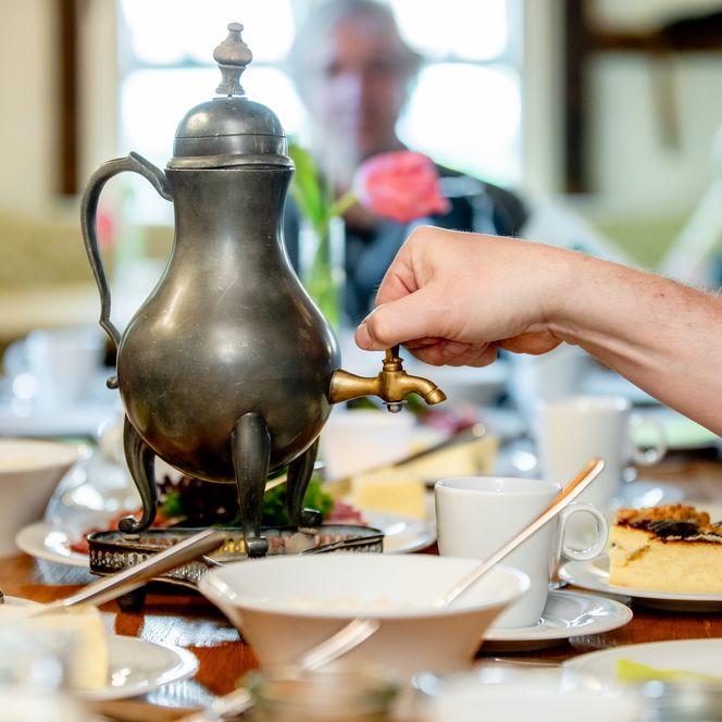 Eine Hand öffnet eine antike Kaffeekanne bei einem gedeckten Tisch mit Kuchen und Tassen.